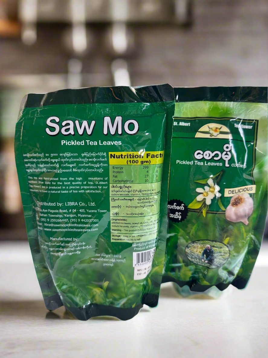 Two packages of Saw Mo Pickled Tea Leaves on a white surface with a blurred background.