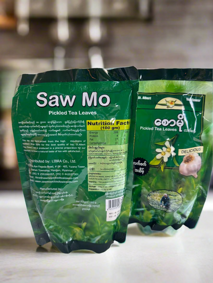 Two packages of Saw Mo Pickled Tea Leaves on a white surface with a blurred background.